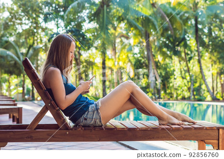Happy smartphone woman relaxing near swimming pool. Beautiful girl using her mobile phone app 4g data to play songs while relaxing on summer luxury vacations Happy smartphone woman relaxing near swimming pool. Beautiful girl using her mobile phone app 4g data to play songs while relaxing on summer luxury vacations 92856760