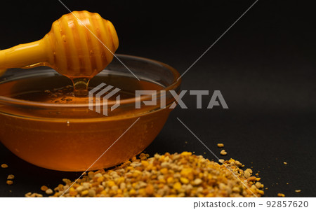 A yellow spoon of honey dipped in a jar of honey and bee bread scattered on a black background. Honey dripping around, nice and inviting photos. Healthy food concept. 92857620