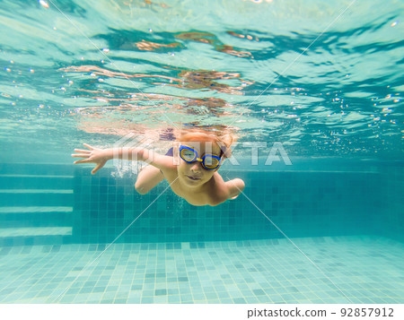 A child boy is swimming underwater in a pool, smiling and holding breath, with swimming glasses 92857912