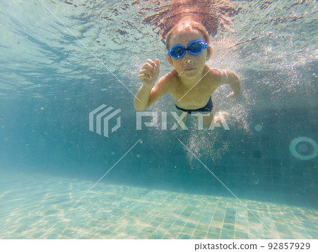 A child boy is swimming underwater in a pool, smiling and holding breath, with swimming glasses 92857929