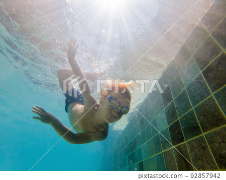 A child boy is swimming underwater in a pool, smiling and holding breath, with swimming glasses 92857942