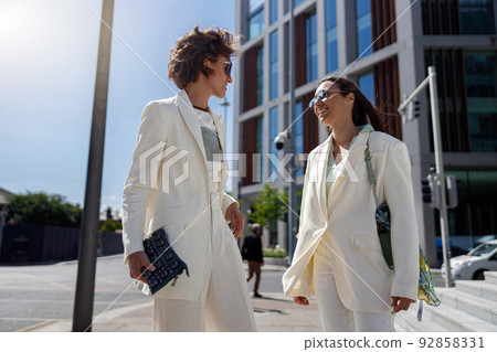Two happy woman in trendy outfits laugh and walk outside on background of modern building Two happy woman in trendy outfits laugh and walk outside on background of modern building 92858331