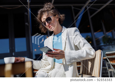 Attractive business woman in sunglasses drinking coffee and using phone sitting in cafe Attractive business woman in sunglasses drinking coffee and using phone sitting in cafe 92858383