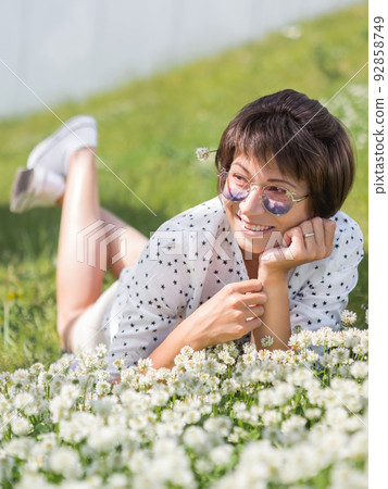 Woman in colorful sunglasses sniffs clover flowers on lawn in urban park. Nature in town. Relax outdoors after work. Summer vibes. Woman in colorful sunglasses sniffs clover flowers on lawn in urban park. Nature in town. Relax outdoors after work. Summer vibes. 92858749