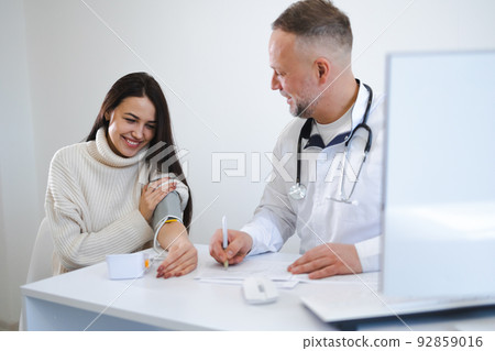 The male doctor measures the woman blood pressure and records the results in the patient's card. Female at the appointment at cardiologist 92859016