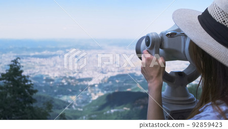 Unrecognizable woman traveler looking through viewing binoculars at summer city. Copy space. Woman in straw hat observes of blur panorama, with binoculars at observation deck. Mountain Dajti Albania Unrecognizable woman traveler looking through viewing binoculars at summer city. Copy space. Woman in straw hat observes of blur panorama, with binoculars at observation deck. Mountain Dajti Albania 92859423