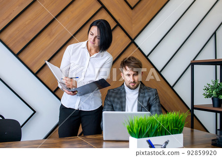 A woman stands next to a man working on a laptop making notes in a folder A woman stands next to a man working on a laptop making notes in a folder 92859920
