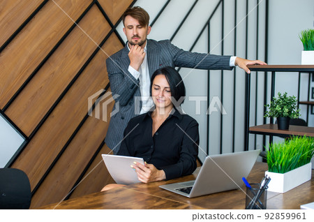 A man with a notebook stands next to a woman sitting at a table with a laptop in the office A man with a notebook stands next to a woman sitting at a table with a laptop in the office 92859961