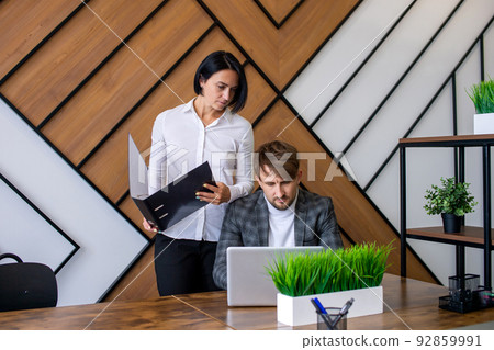 A woman in a white shirt with an open folder is standing next to a man sitting at a laptop 92859991