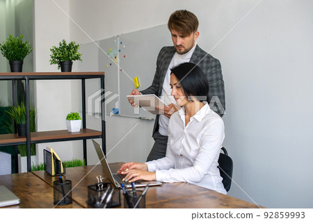 A man with a notebook stands next to a woman sitting at a table with a laptop in the office 92859993