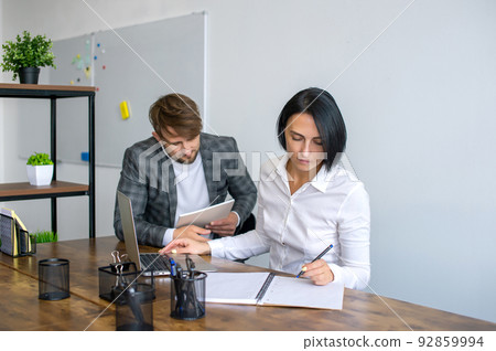 A man with a notebook stands next to a woman sitting at a table with a laptop in the office 92859994