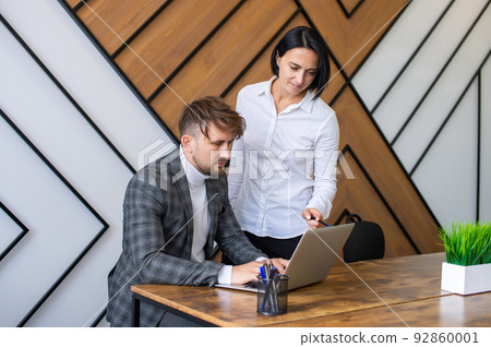 A female supervisor stands next to a male employee working at a laptop 92860001