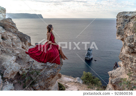A girl with loose hair in a red dress sits on a rock rock above the sea. In the background, the sea. The concept of travel. 92860036