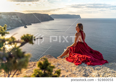 A girl with loose hair in a red dress sits on a rock rock above the sea. In the background, the sun rises from behind the mountains. 92860058
