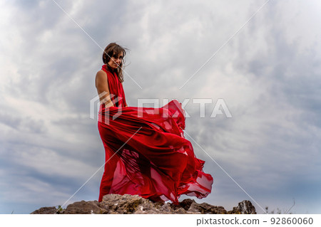 A woman in a red dress stands above a stormy sky, her dress fluttering, the fabric flying in the wind. A woman in a red dress stands above a stormy sky, her dress fluttering, the fabric flying in the wind. 92860060