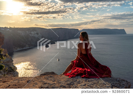 A girl with loose hair in a red dress sits on a rock rock above the sea. In the background, the sun rises from behind the mountains. 92860071