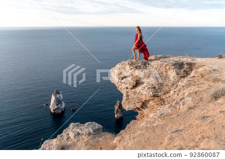 A girl with loose hair in a red dress stands on a rock rock above the sea. In the background, the sea and the rocks. The concept of travel 92860087