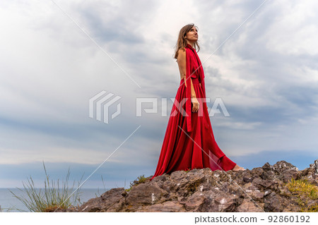 A woman in a red dress stands above a stormy sky, her dress fluttering, the fabric flying in the wind. 92860192