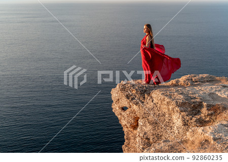 A girl with loose hair in a red dress stands on a rock rock above the sea. In the background, the sea and the rocks. The concept of travel 92860235