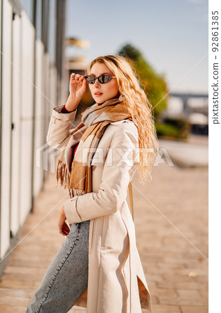 student or businesswoman in sunglasses and a coat with scarf walks down a street 92861385
