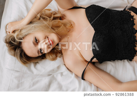 a blonde woman in black underwear on a bed with white linens.  92861388