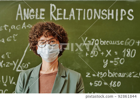 Young teacher in eyeglasses and protective mask standing by blackboard Young teacher in eyeglasses and protective mask standing by blackboard 92861844