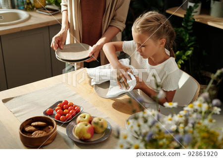 Cute blond youthful girl with white duster wiping clean plates after washing 92861920