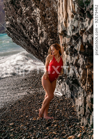 A beauty in a red swimsuit with long legs poses on a fantastic beach with huge waves against the background of mountains 92879462
