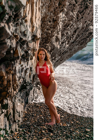 A beauty in a red swimsuit with long legs poses on a fantastic beach with huge waves against the background of mountains 92879466