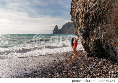 A beauty in a red swimsuit with long legs poses on a fantastic beach with huge waves against the background of mountains 92879525