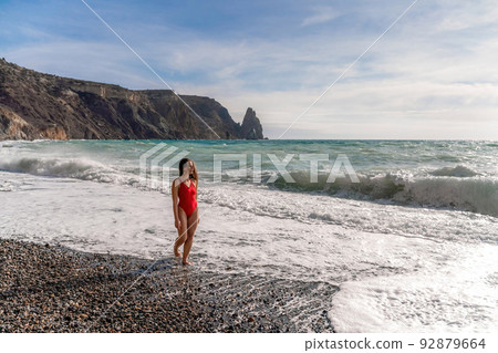 A beautiful and sexy brunette in a red swimsuit on a pebble beach, Running along the shore in the foam of the waves 92879664
