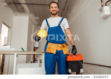 Hard working contractor, handyman in uniform with toolbox smiling at camera while standing indoors Hard working contractor, handyman in uniform with toolbox smiling at camera while standing indoors 92880337