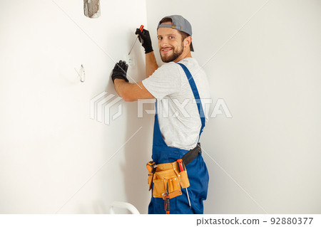 Enthusiastic electrician, handyman looking at camera with a smile while working on wiring in new house, standing on a ladder Enthusiastic electrician, handyman looking at camera with a smile while working on wiring in new house, standing on a ladder 92880377