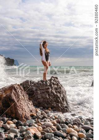 A beautiful girl in a white shirt and black swimsuit stands on a rock, big waves with white foam. A cloudy stormy day at sea, with clouds and big waves hitting the rocks. 92880460