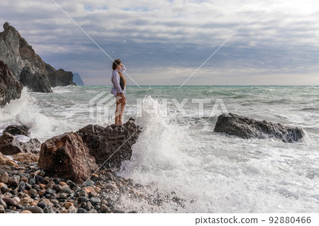 A beautiful girl in a white shirt and black swimsuit stands on a rock, big waves with white foam. A cloudy stormy day at sea, with clouds and big waves hitting the rocks. 92880466