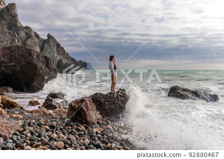 A beautiful girl in a white shirt and black swimsuit stands on a rock, big waves with white foam. A cloudy stormy day at sea, with clouds and big waves hitting the rocks. 92880467