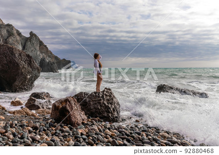 A beautiful girl in a white shirt and black swimsuit stands on a rock, big waves with white foam. A cloudy stormy day at sea, with clouds and big waves hitting the rocks. 92880468