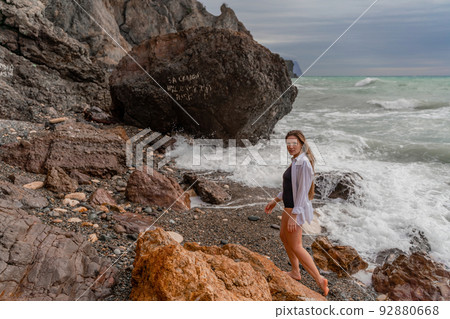 A beautiful girl in a black dress is walking on the waves, big waves with white foam. A cloudy stormy day at sea, with clouds and big waves hitting the rocks. 92880668