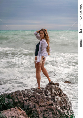 A beautiful girl in a white shirt and black swimsuit stands on a rock, big waves with white foam. A cloudy stormy day at sea, with clouds and big waves hitting the rocks. 92880669