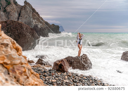 A beautiful girl in a white shirt and black swimsuit stands on a rock, big waves with white foam. A cloudy stormy day at sea, with clouds and big waves hitting the rocks. A beautiful girl in a white shirt and black swimsuit stands on a rock, big waves with white foam. A cloudy stormy day at sea, with clouds and big waves hitting the rocks. 92880675