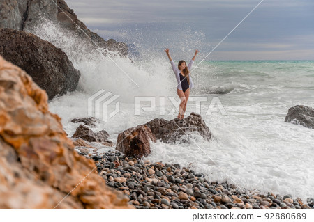 A beautiful girl in a white shirt and black swimsuit stands on a rock, big waves with white foam. A cloudy stormy day at sea, with clouds and big waves hitting the rocks. 92880689