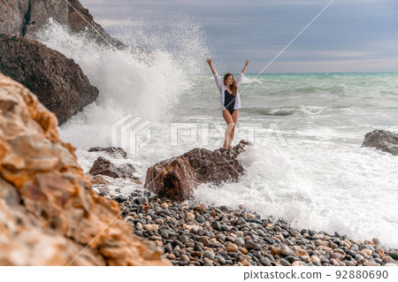 A beautiful girl in a white shirt and black swimsuit stands on a rock, big waves with white foam. A cloudy stormy day at sea, with clouds and big waves hitting the rocks. 92880690