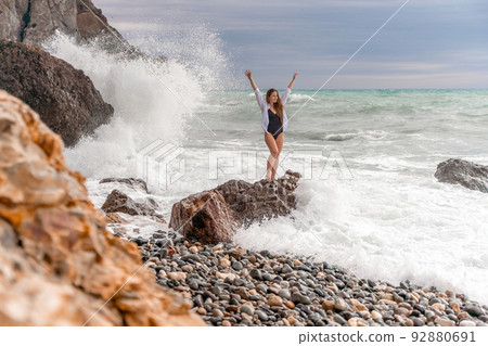 A beautiful girl in a white shirt and black swimsuit stands on a rock, big waves with white foam. A cloudy stormy day at sea, with clouds and big waves hitting the rocks. 92880691