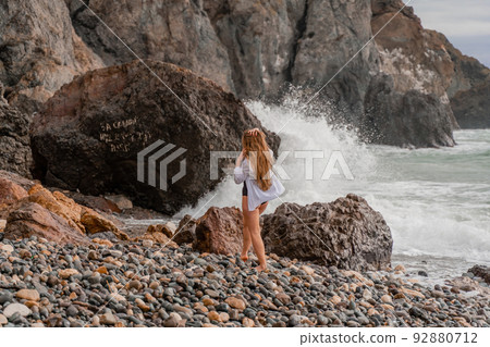 A beautiful girl in a white shirt and black swimsuit stands on the edge of a cliff, big waves with white foam. A cloudy stormy day at sea, with clouds and big waves hitting the rocks. A beautiful girl in a white shirt and black swimsuit stands on the edge of a cliff, big waves with white foam. A cloudy stormy day at sea, with clouds and big waves hitting the rocks. 92880712