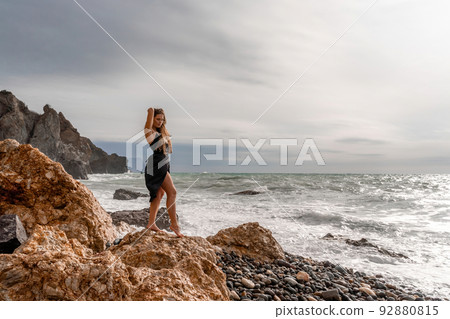A beautiful girl in a black dress stands on a rock, big waves with white foam. A cloudy stormy day at sea, with clouds and big waves hitting the rocks. 92880815