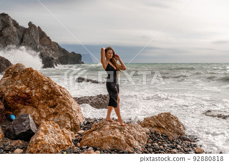 A beautiful girl in a black dress stands on a rock, big waves with white foam. A cloudy stormy day at sea, with clouds and big waves hitting the rocks. 92880818