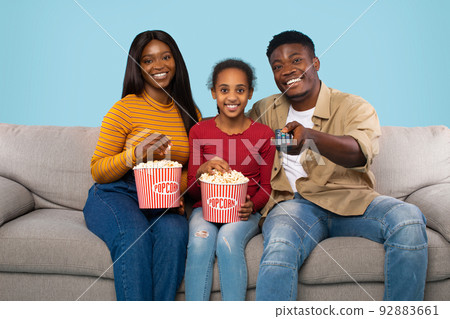 Weekend leisure. Black family watching TV and eating popcorn, sitting on couch over blue studio background 92883661