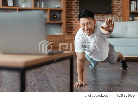 Online sports. Sporty asian mature man doing exercises near laptop, standing in plank pose and raising one hand Online sports. Sporty asian mature man doing exercises near laptop, standing in plank pose and raising one hand 92883690