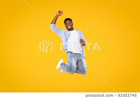 Carefree excited black man in casual outfit jumping in the air and smiling over yellow studio background, copy space 92883748