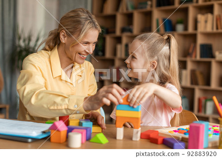 Cheerful child development specialist having session with cute little girl, playing with colorful wooden bricks Cheerful child development specialist having session with cute little girl, playing with colorful wooden bricks 92883920